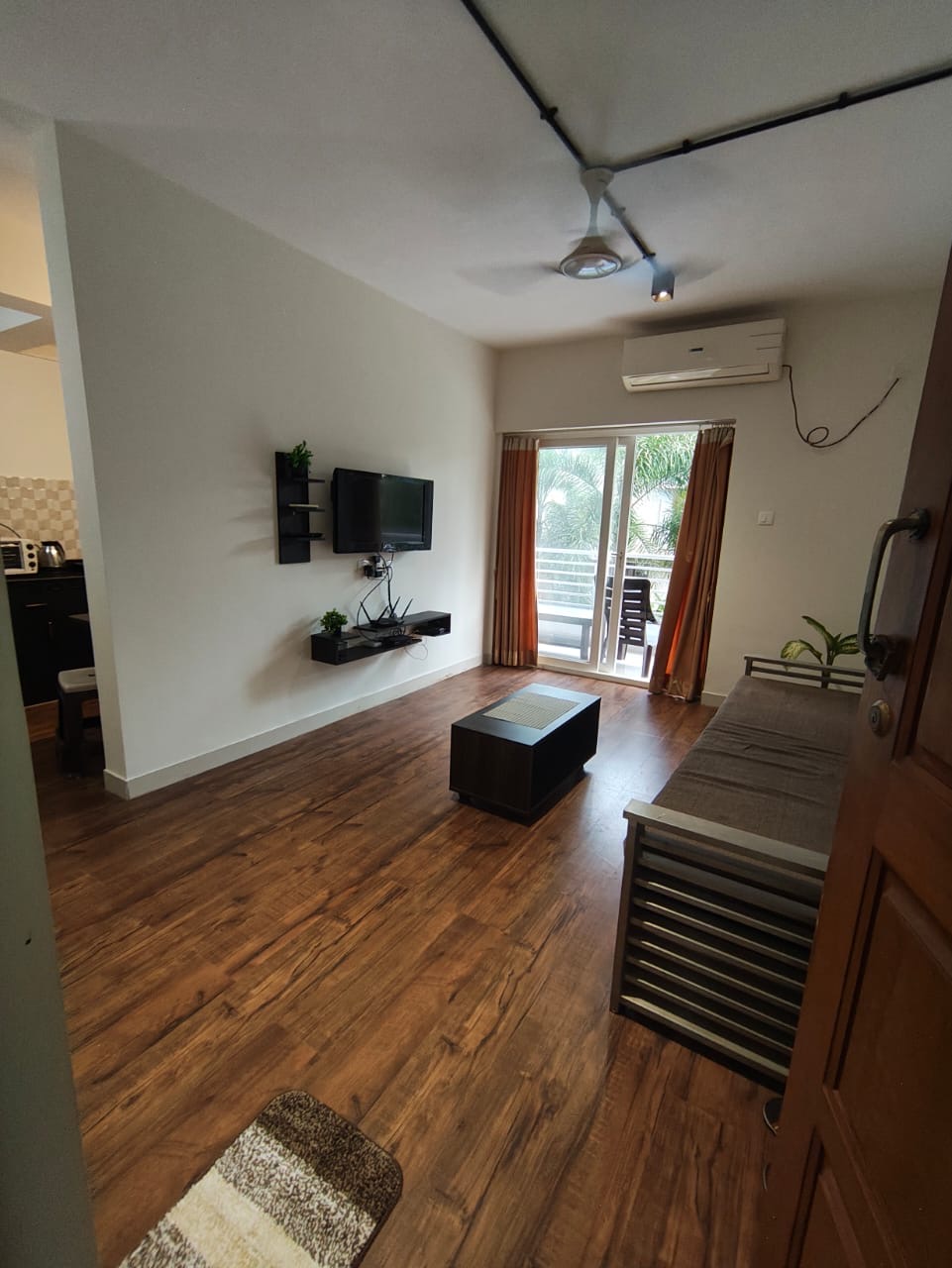 Living room with wall-mounted TV, daybed sofa, and balcony opening to palm-tree views
